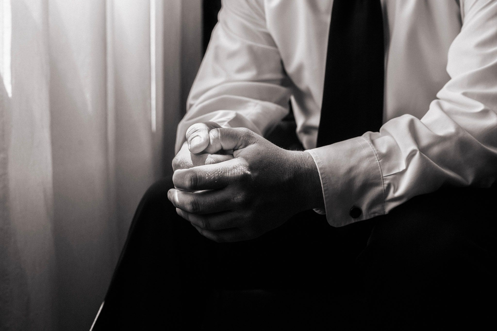 Black and white close-up of a groom in a white shirt and tie clasping his hands nervously.