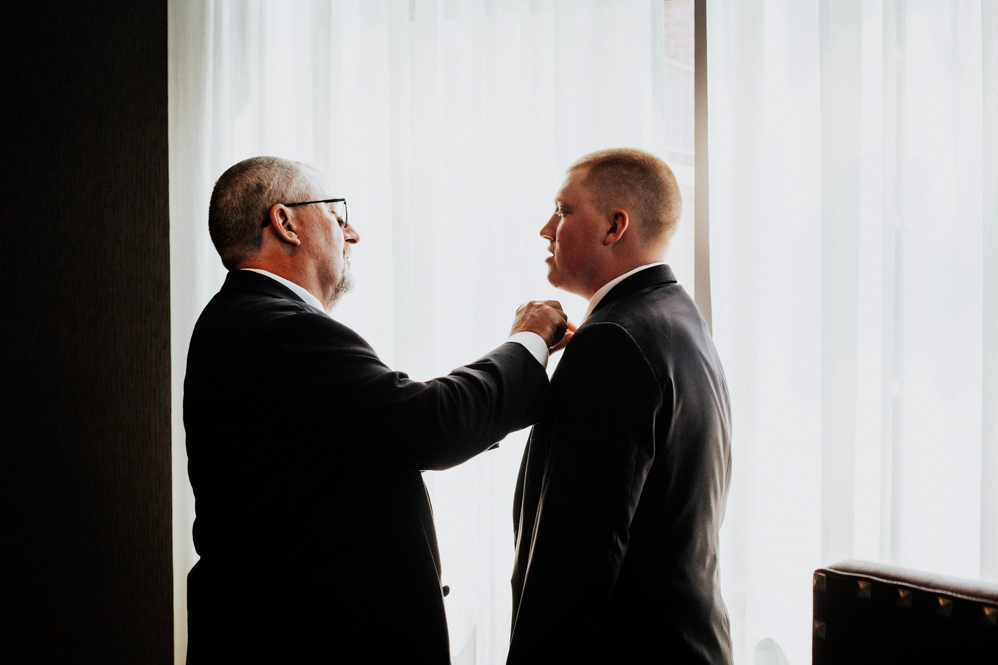 An older man adjusts the bow tie of a younger man in suits, sharing a tender moment by a bright window.