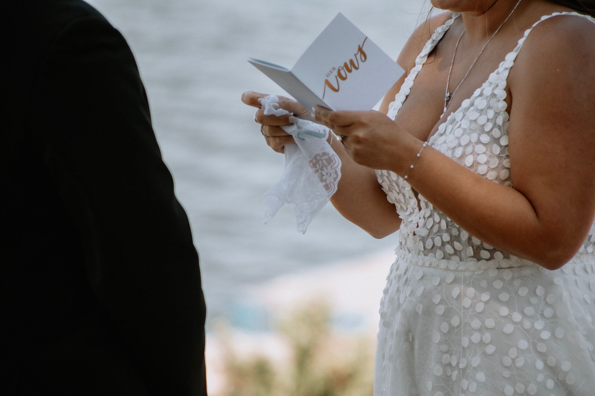 Bride reading wedding vows from a small card during an outdoor ceremony near the water.