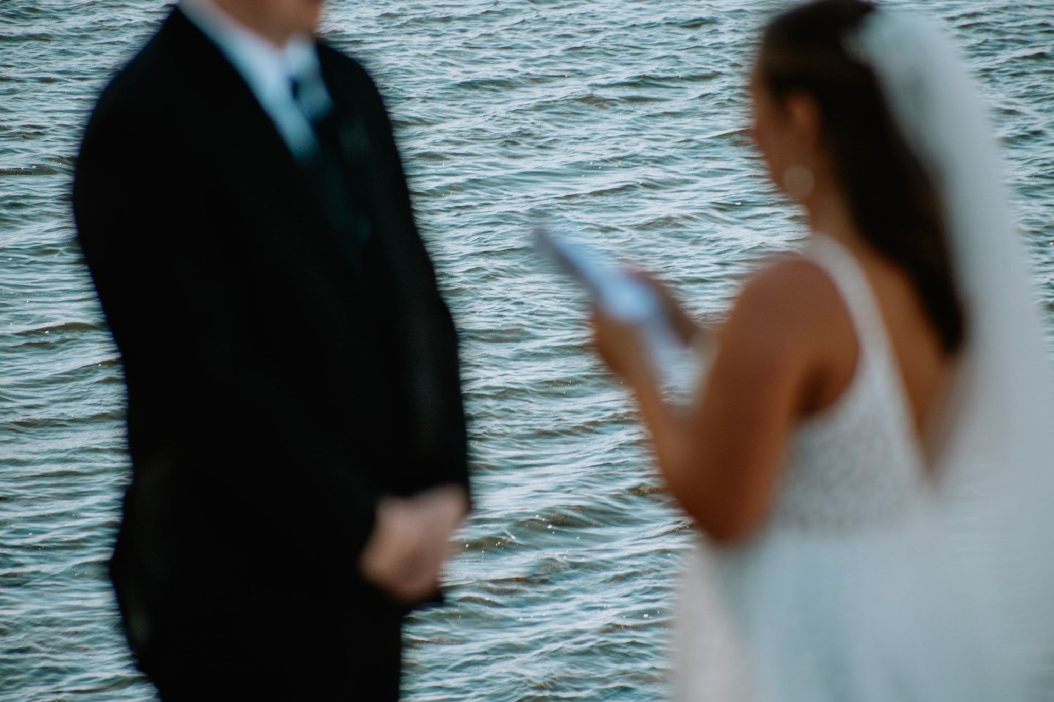 A blurred artistic photo of a bride reading vows to a groom standing near a shimmering body of water.