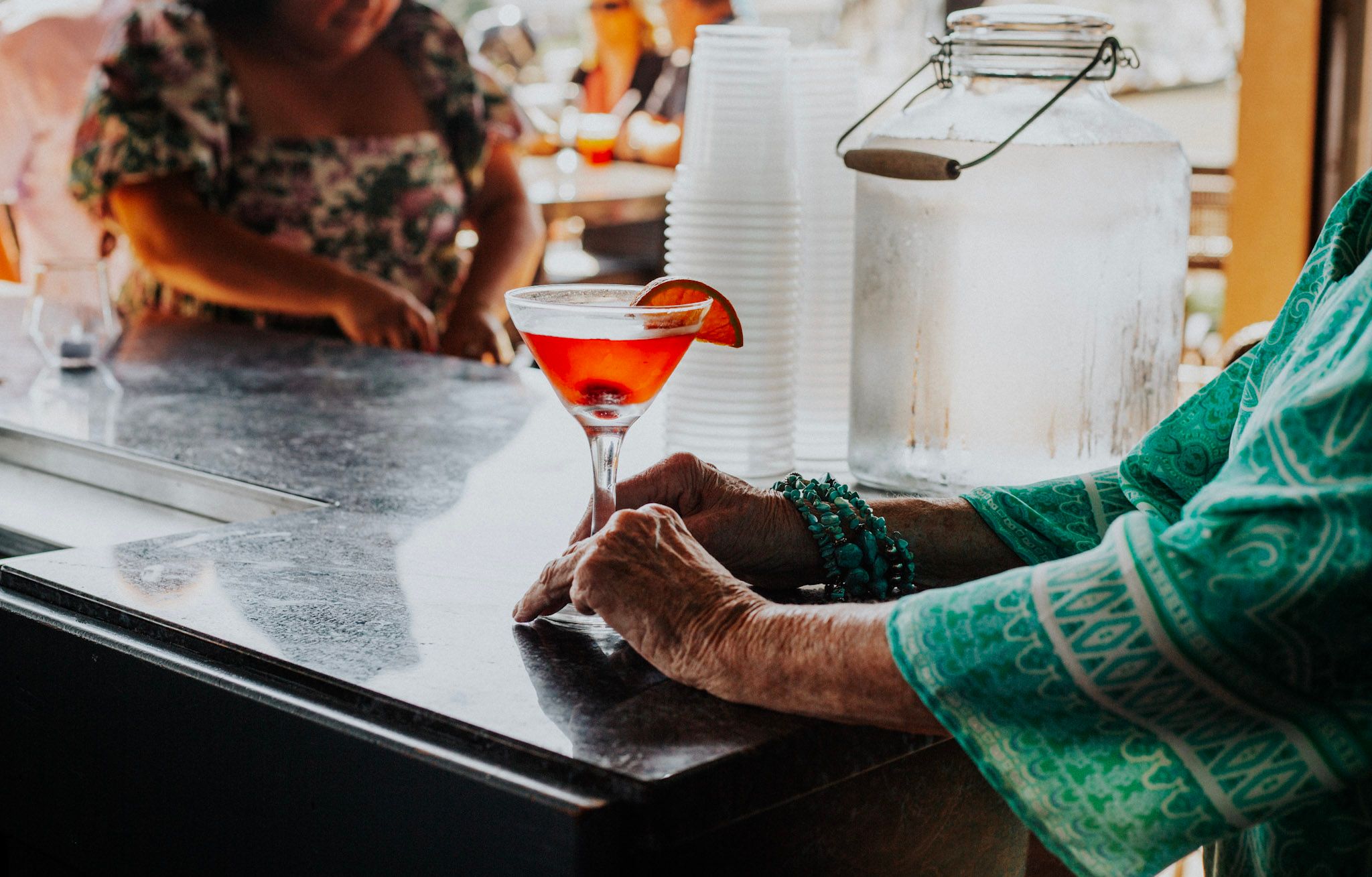 An elderly woman in teal holds a vibrant red cocktail in a martini glass at an outdoor bar.