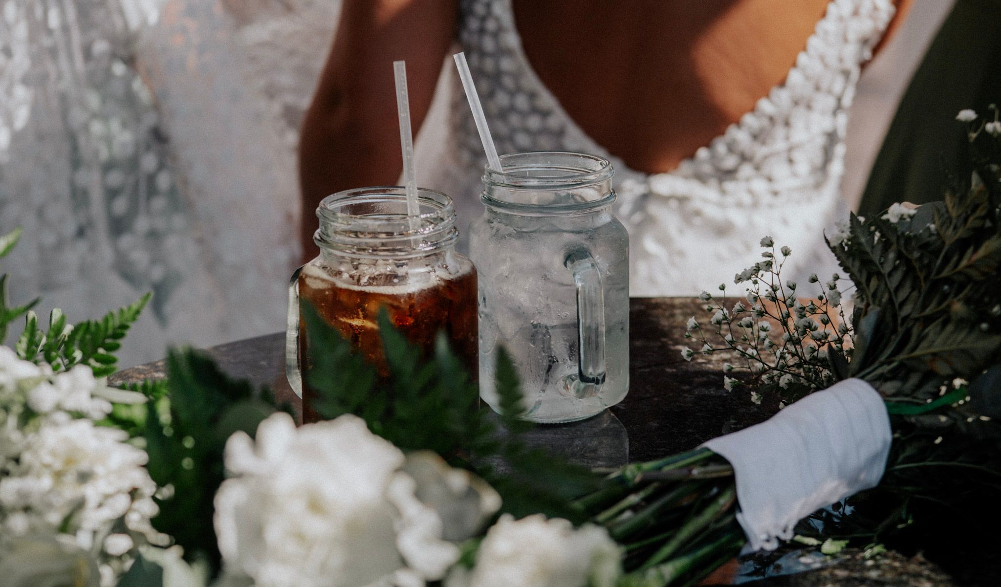 Two mason jar drinks with metal straws sit on a wooden tray surrounded by white wedding flowers.
