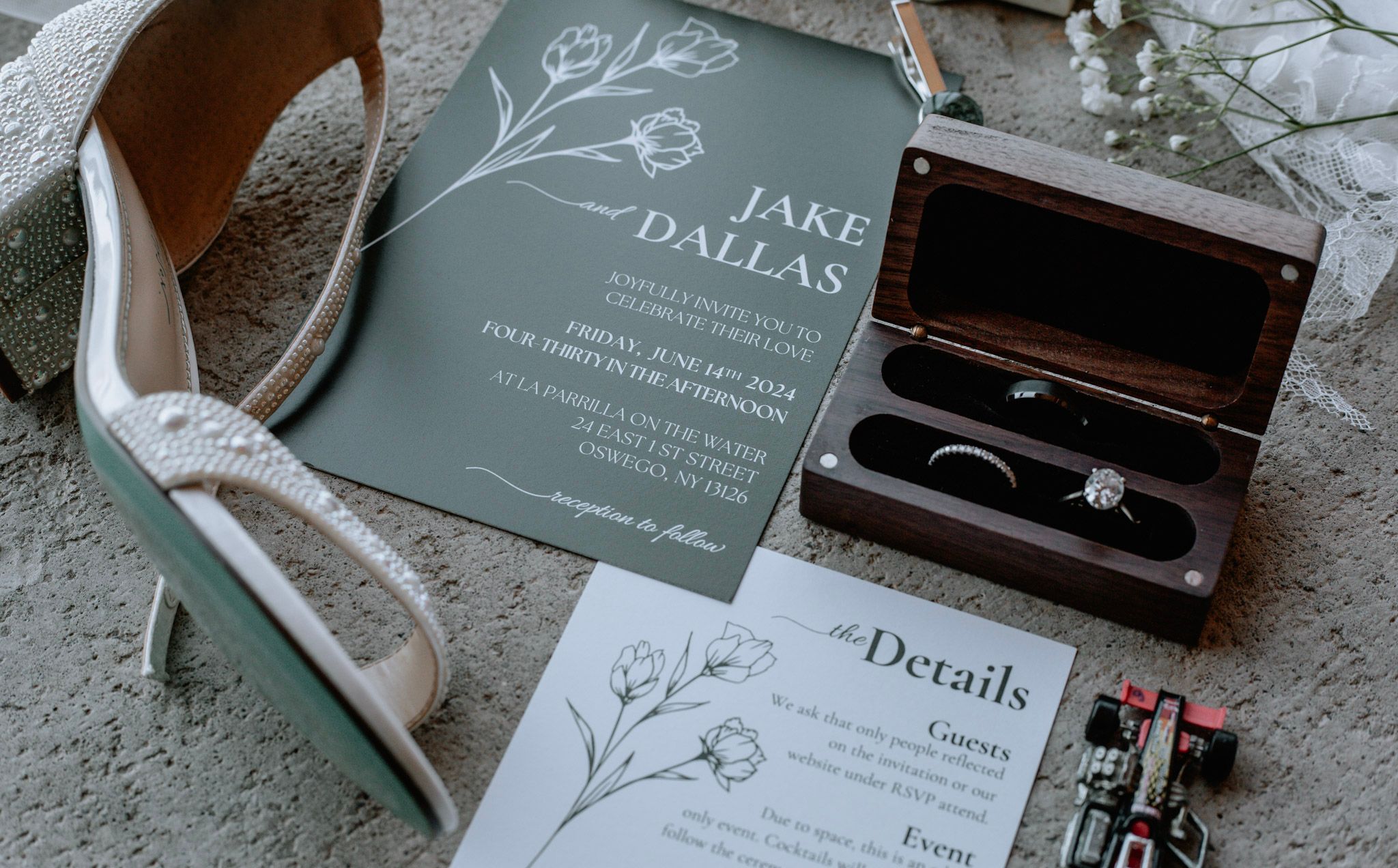 Wedding flat lay featuring sage green invitation, diamond ring in wooden box, and heels.