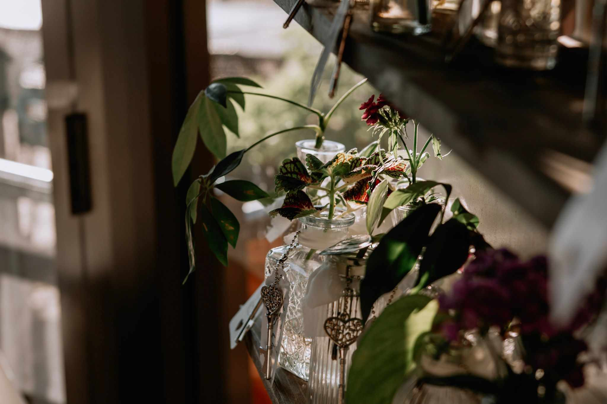 A glass vase with tropical green leaves and small flowers sits on a shelf near a sunlit window.