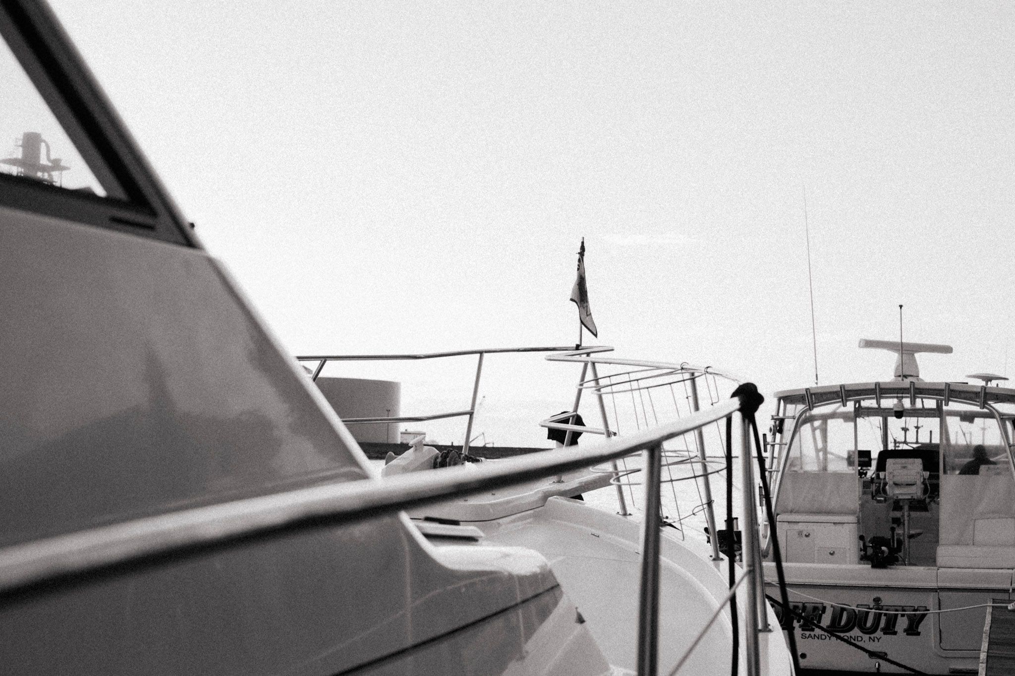Black and white photo of luxury yachts docked at a marina with a flag visible in the background.