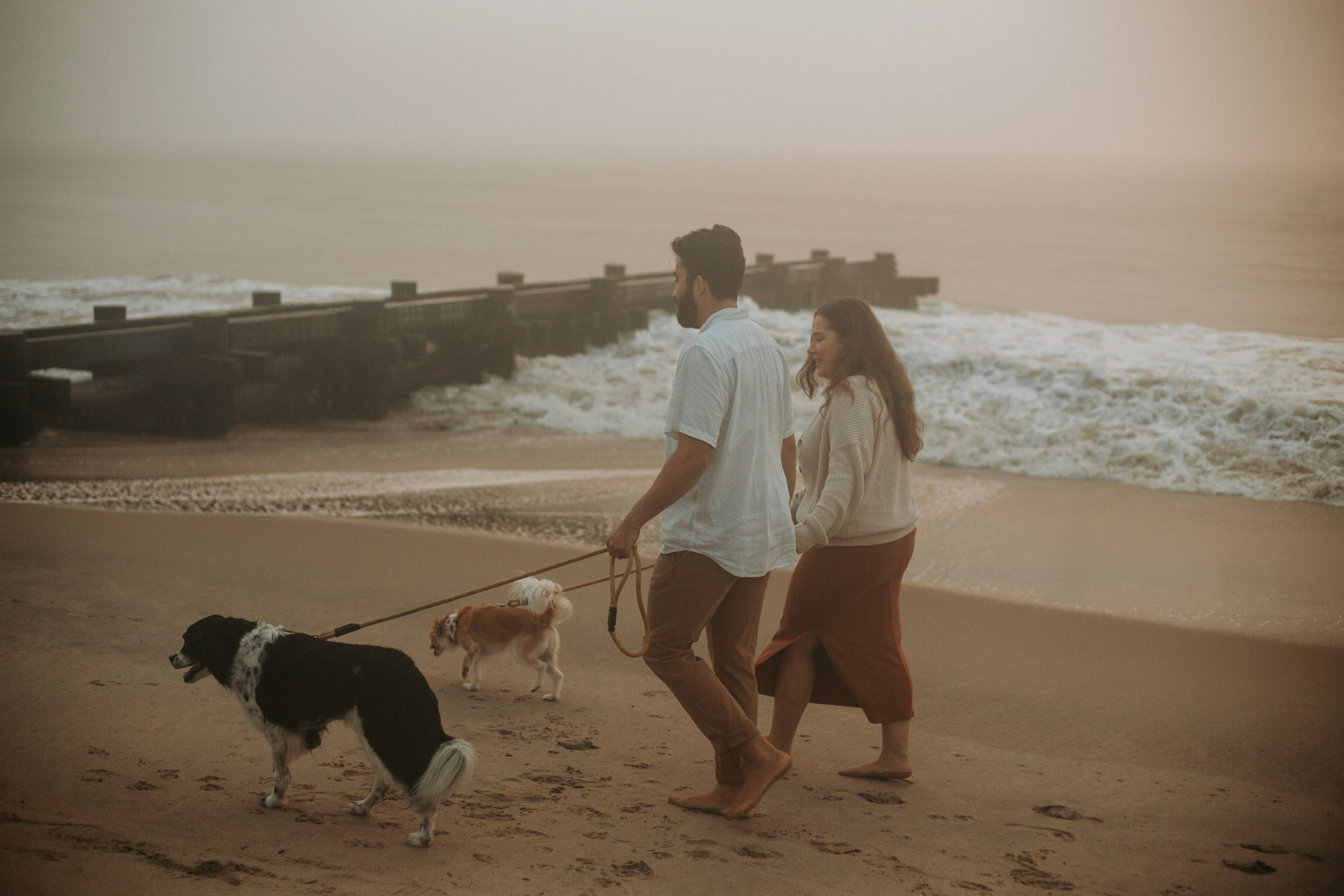 A couple walks two dogs along a misty beach at sunset, enjoying a romantic seaside stroll.