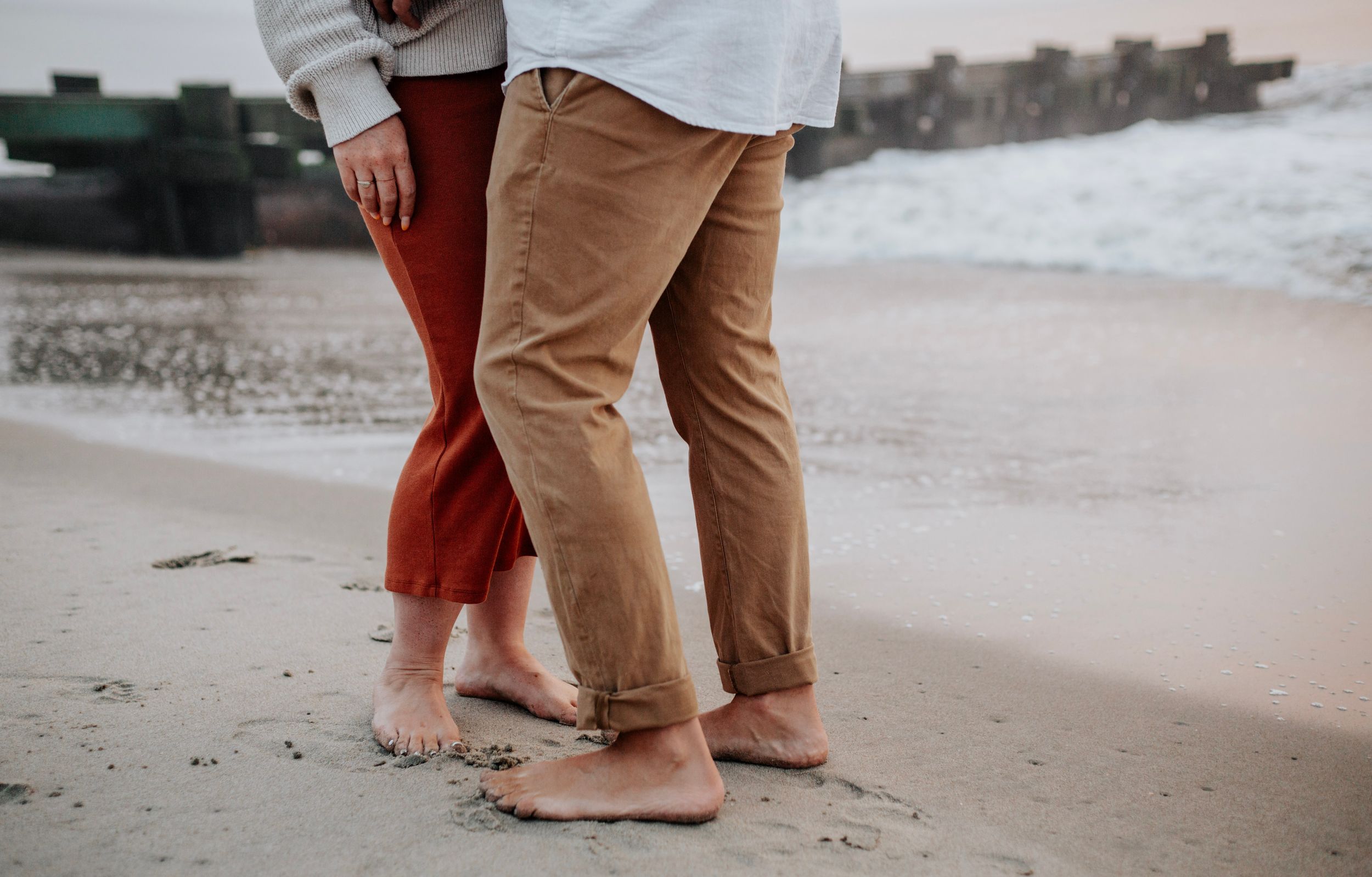 Barefoot couple standing close together on sandy beach with ocean waves in the background.