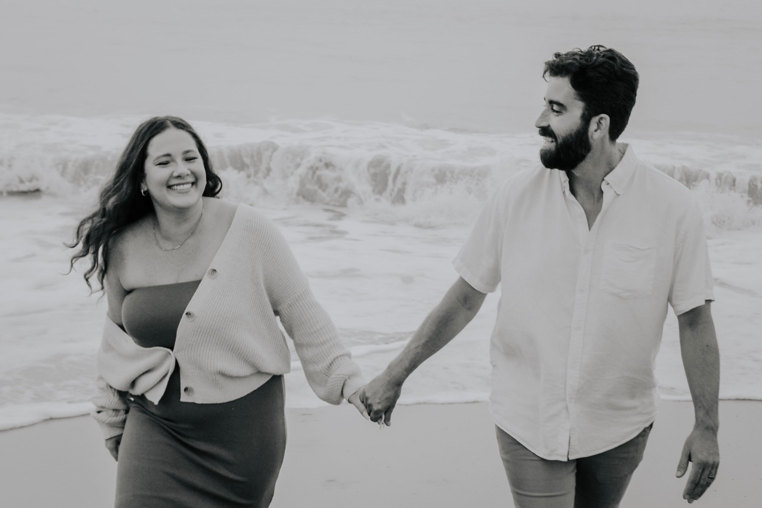 Happy couple holding hands and walking along a beach with ocean waves in black and white.