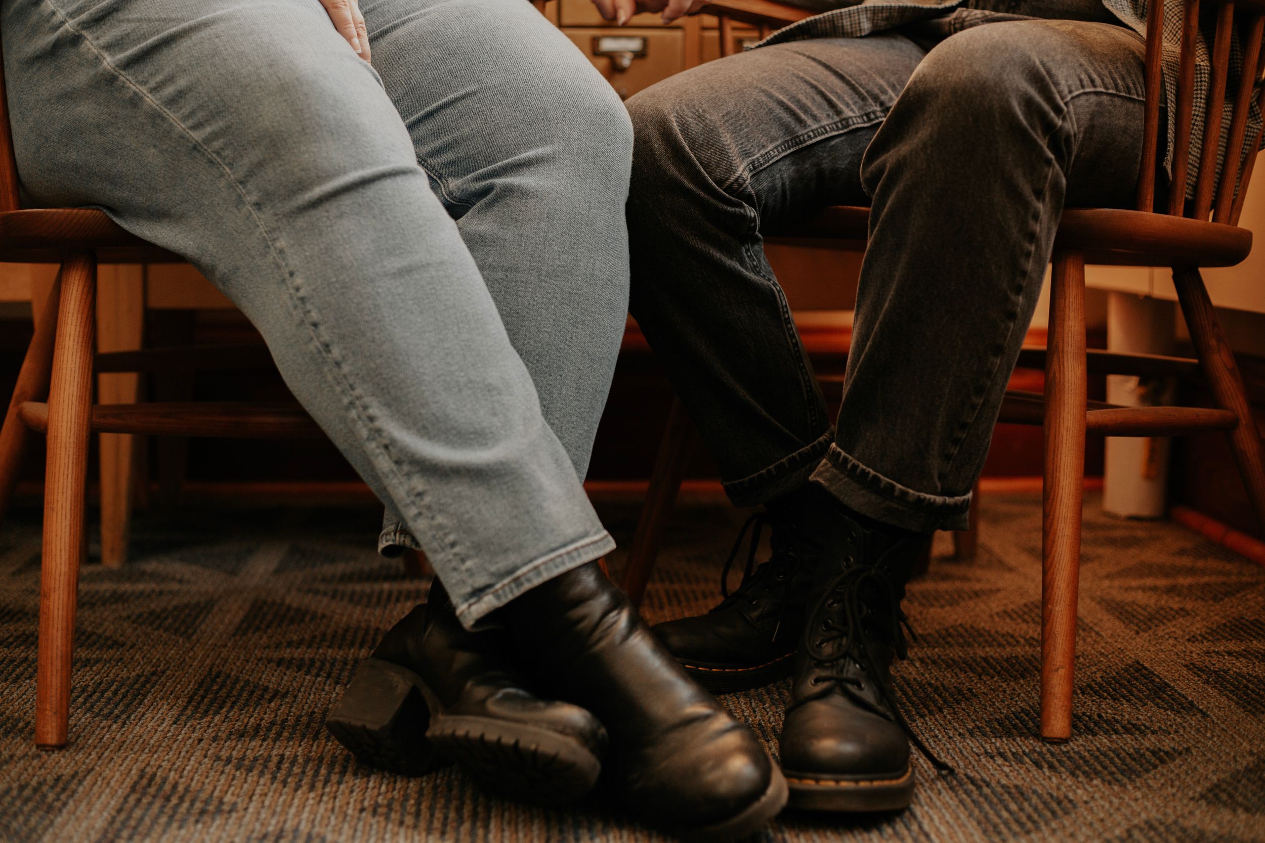 Two people sitting in chairs wearing jeans and black leather boots on a patterned carpet floor.