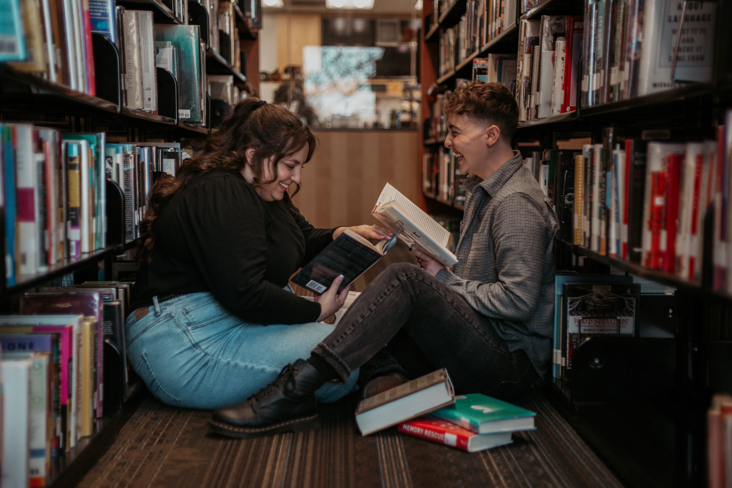 Two people sitting on a library floor between bookshelves, laughing and reading books together.