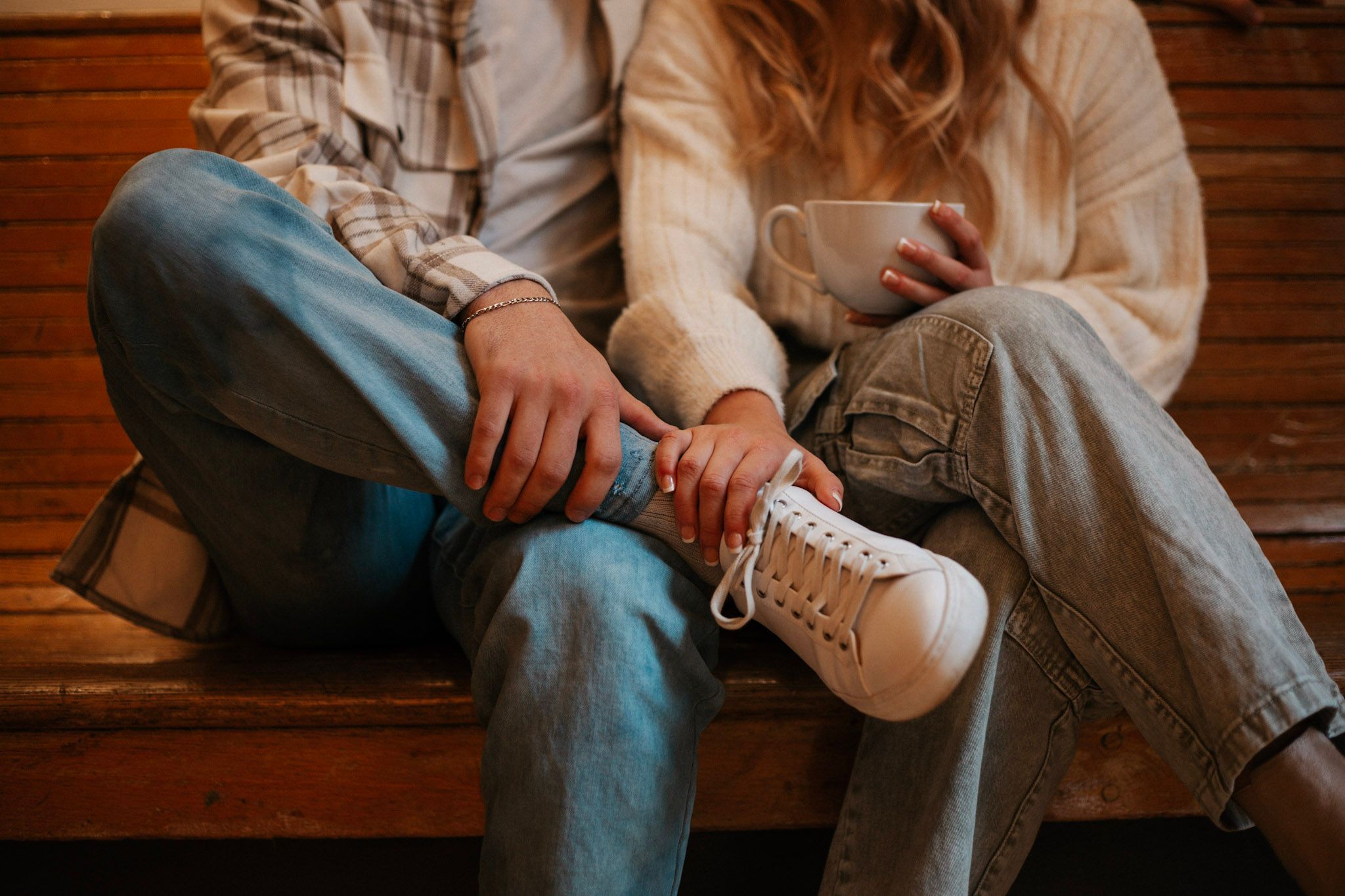 Couple sitting on a wooden bench, one person helping tie white sneaker laces.