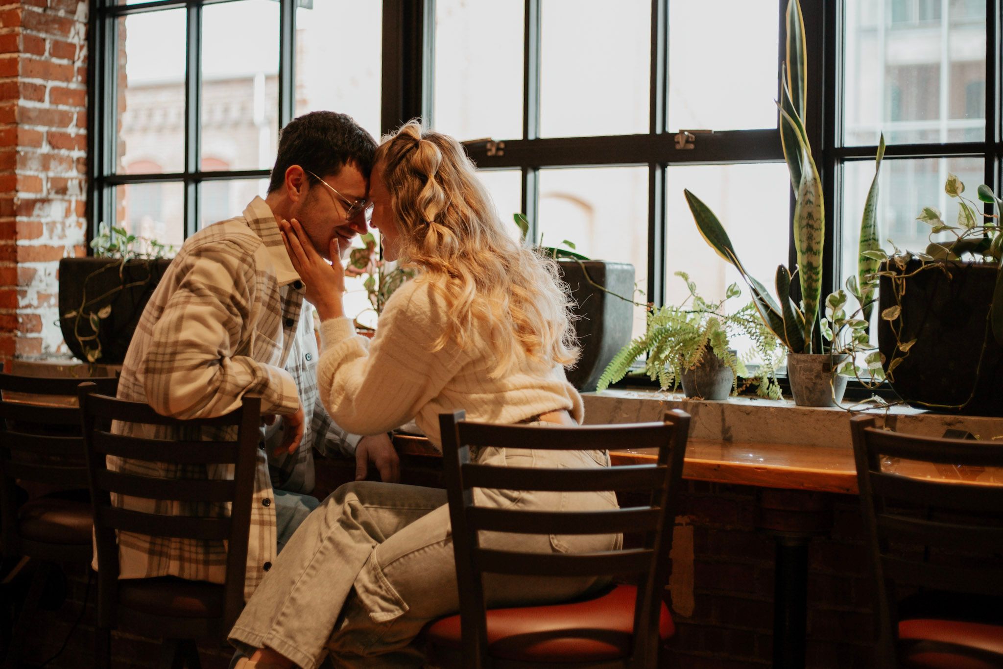 Romantic couple sharing an intimate moment at a cafe table near large windows with plants.