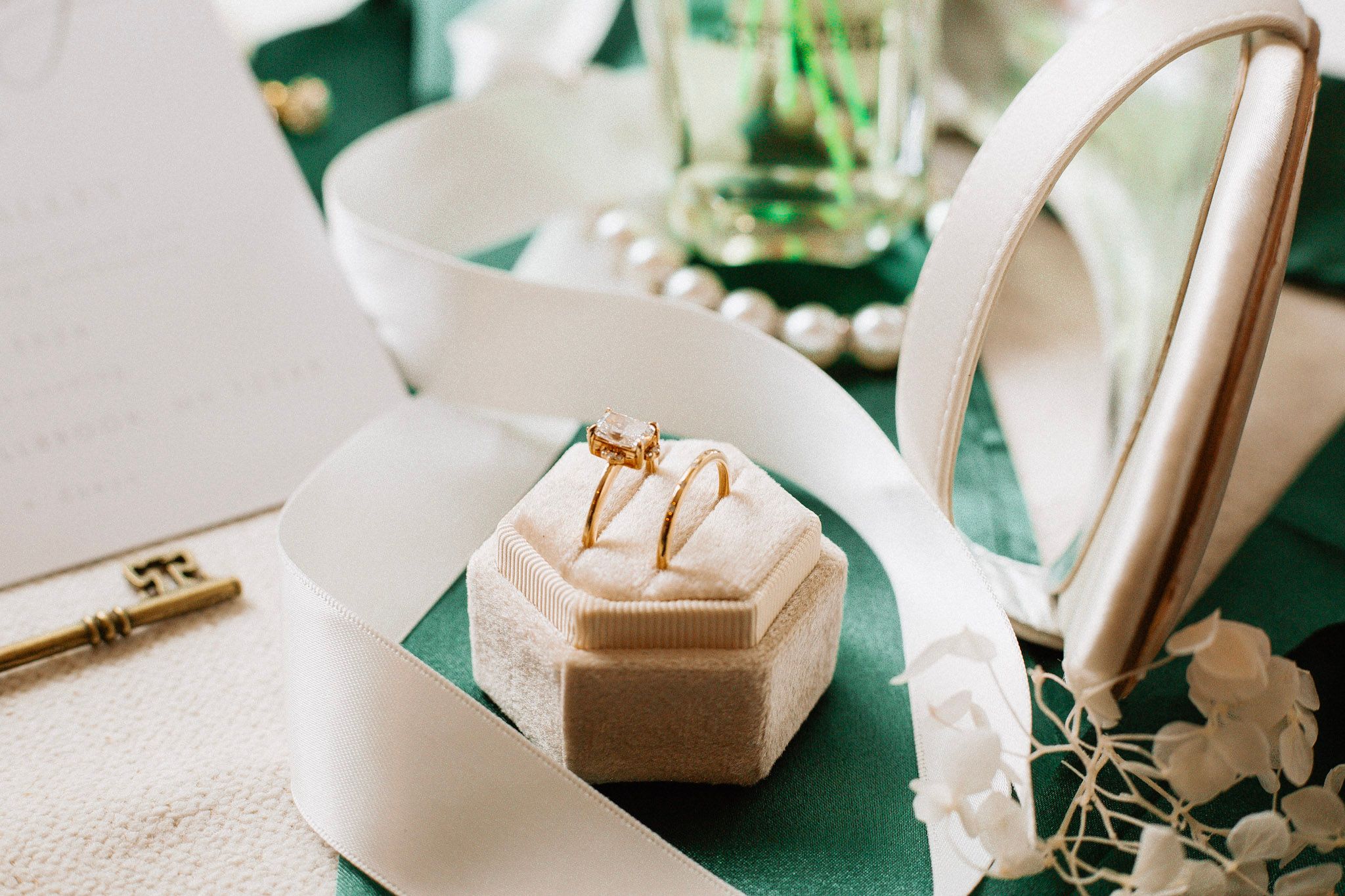 A gold diamond engagement ring in a velvet box rests on a white ribbon on a green velvet surface.