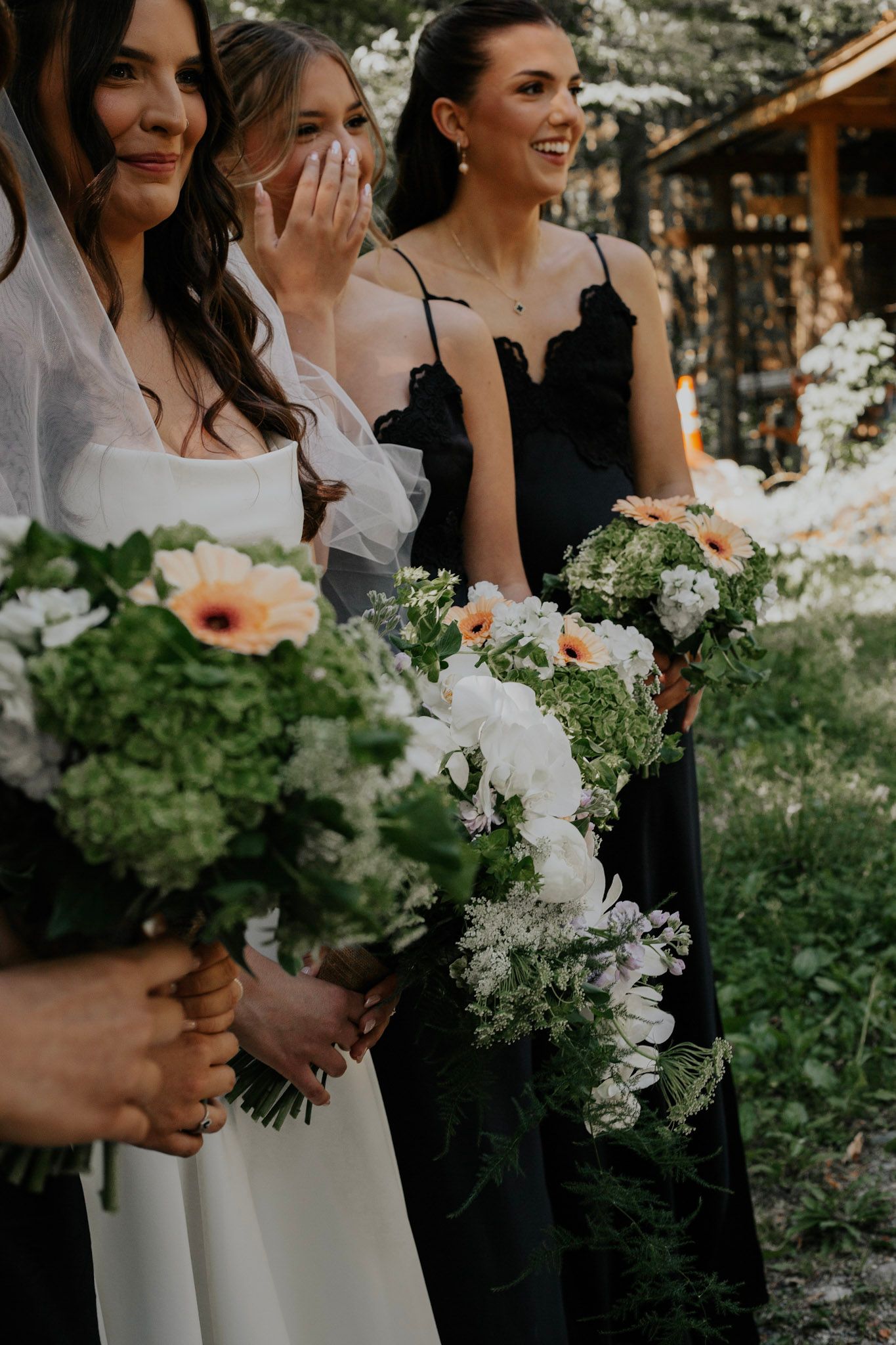Bride and bridesmaids in black dresses holding bouquets of white flowers and greenery outdoors.