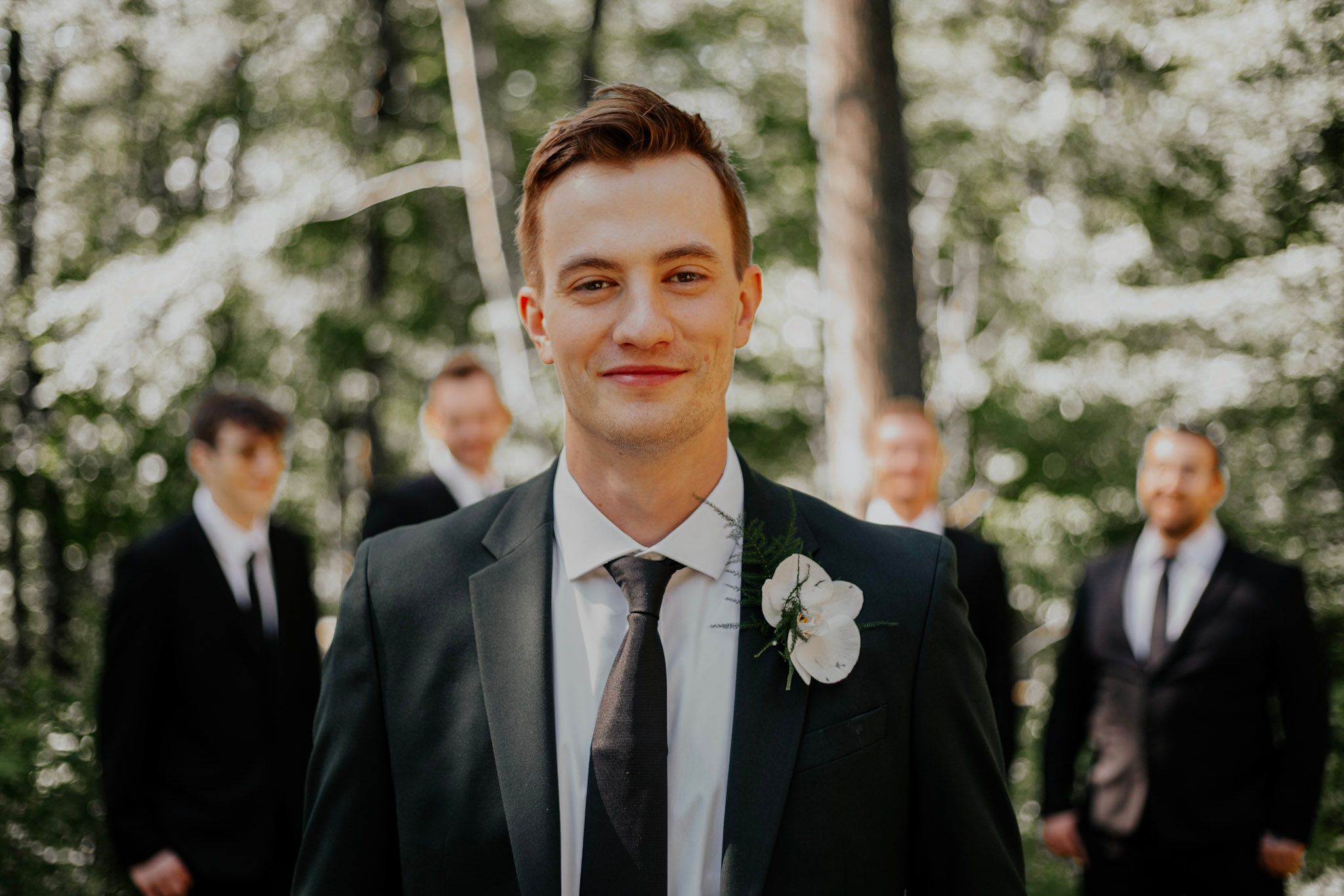 Smiling groom in dark suit with white boutonniere standing before groomsmen in forest.