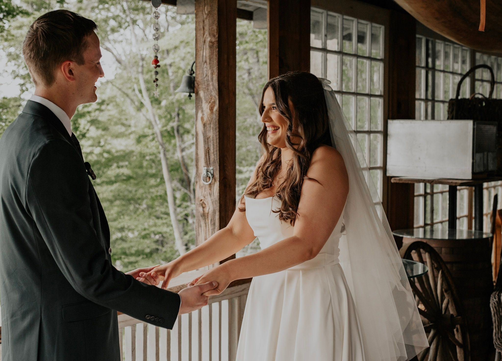 A smiling bride in a white gown and veil holds hands with a groom on a wooden porch surrounded by trees.