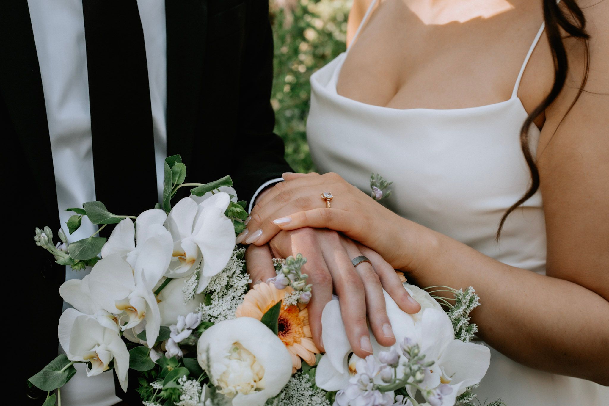 Bride and groom holding hands over a white orchid wedding bouquet, showing wedding rings.