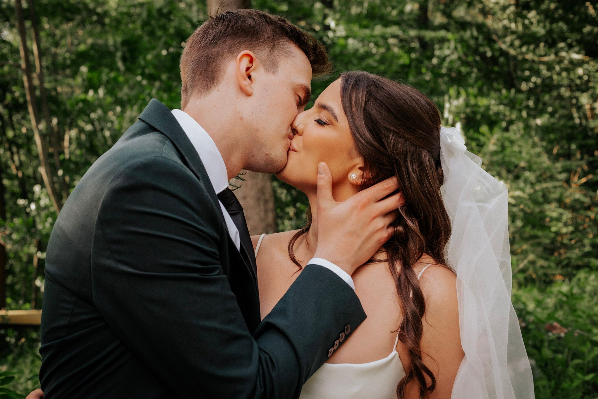 A bride and groom share a romantic first kiss surrounded by lush green trees on their wedding day.