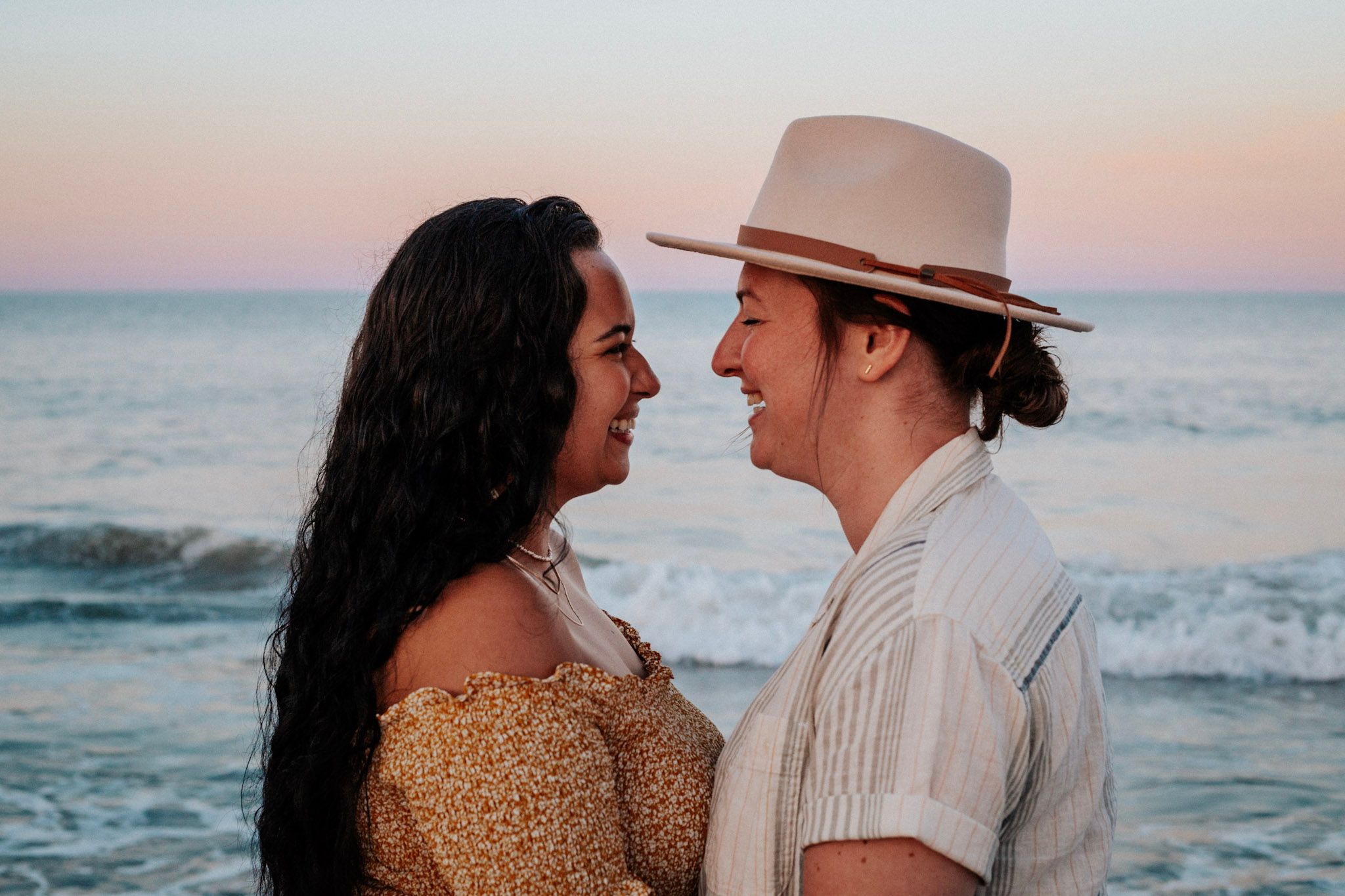 A couple smiles at each other on a beach at sunset, woman in gold dress and man in white linen outfit.