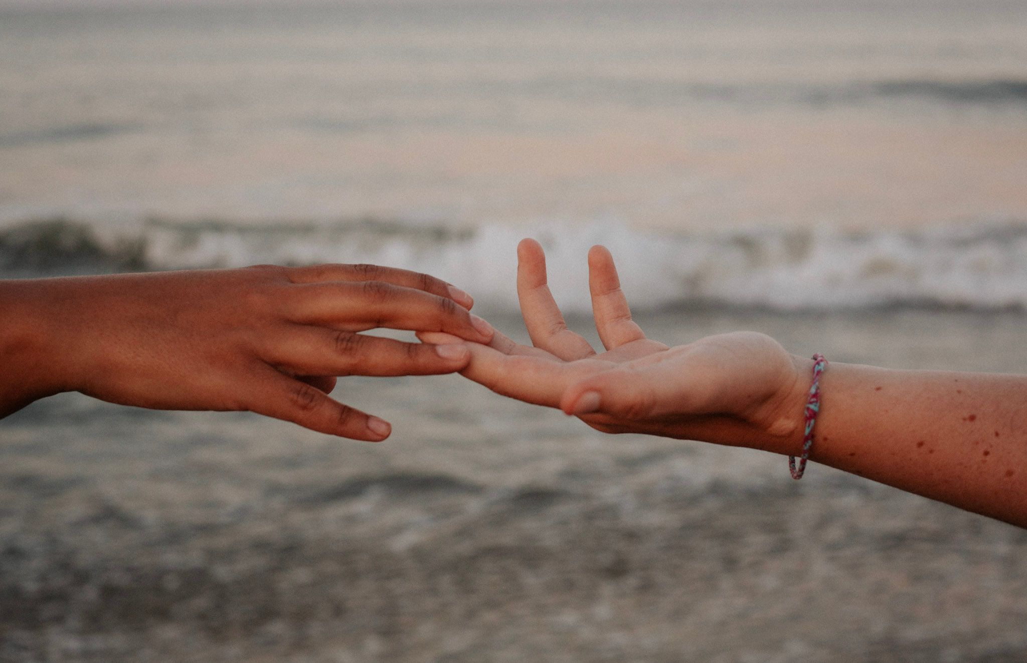 Two hands reaching toward each other on a beach at sunset, one wearing a delicate bracelet.