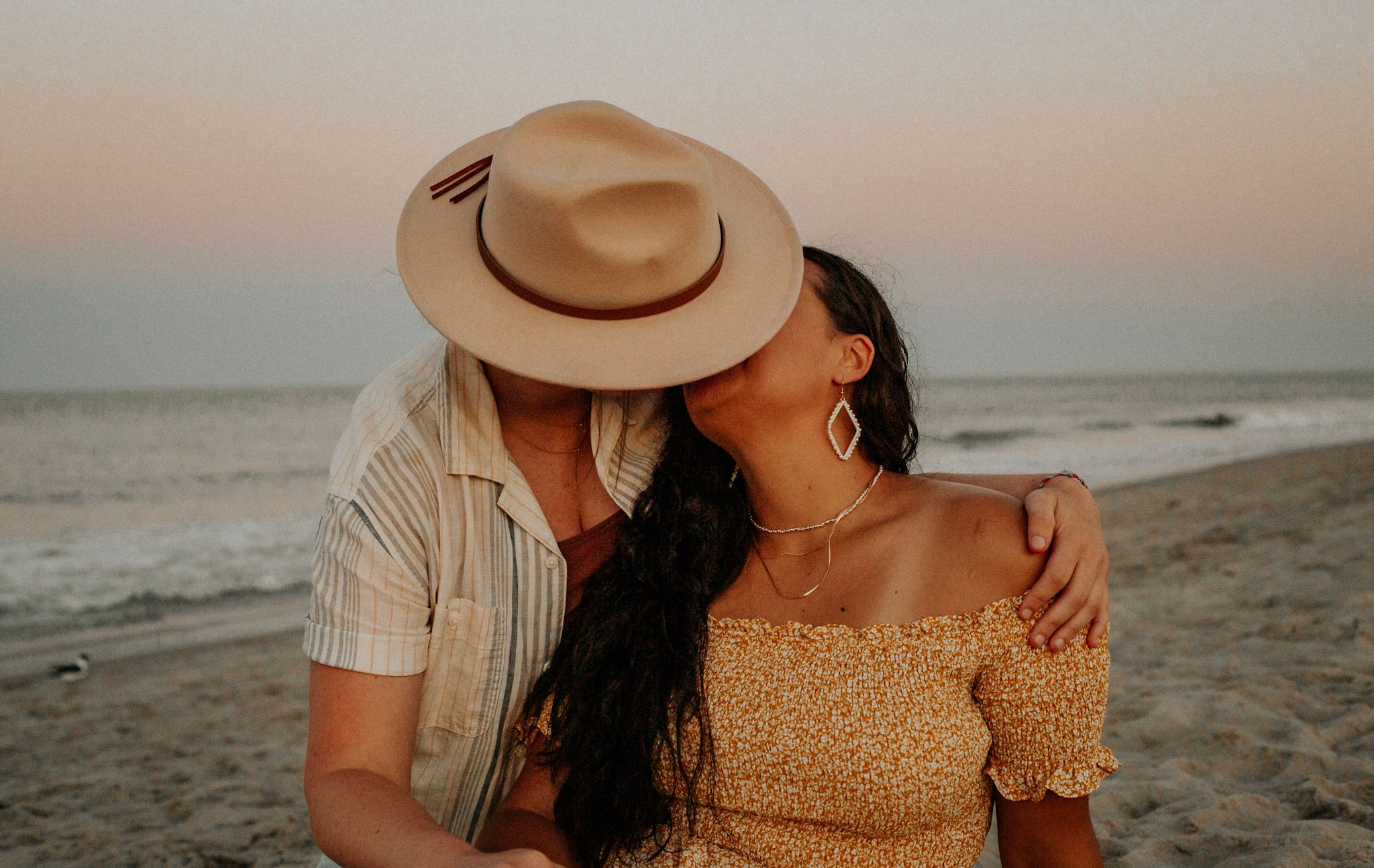 Couple sharing a romantic kiss on the beach at sunset, man wearing a tan fedora hat.