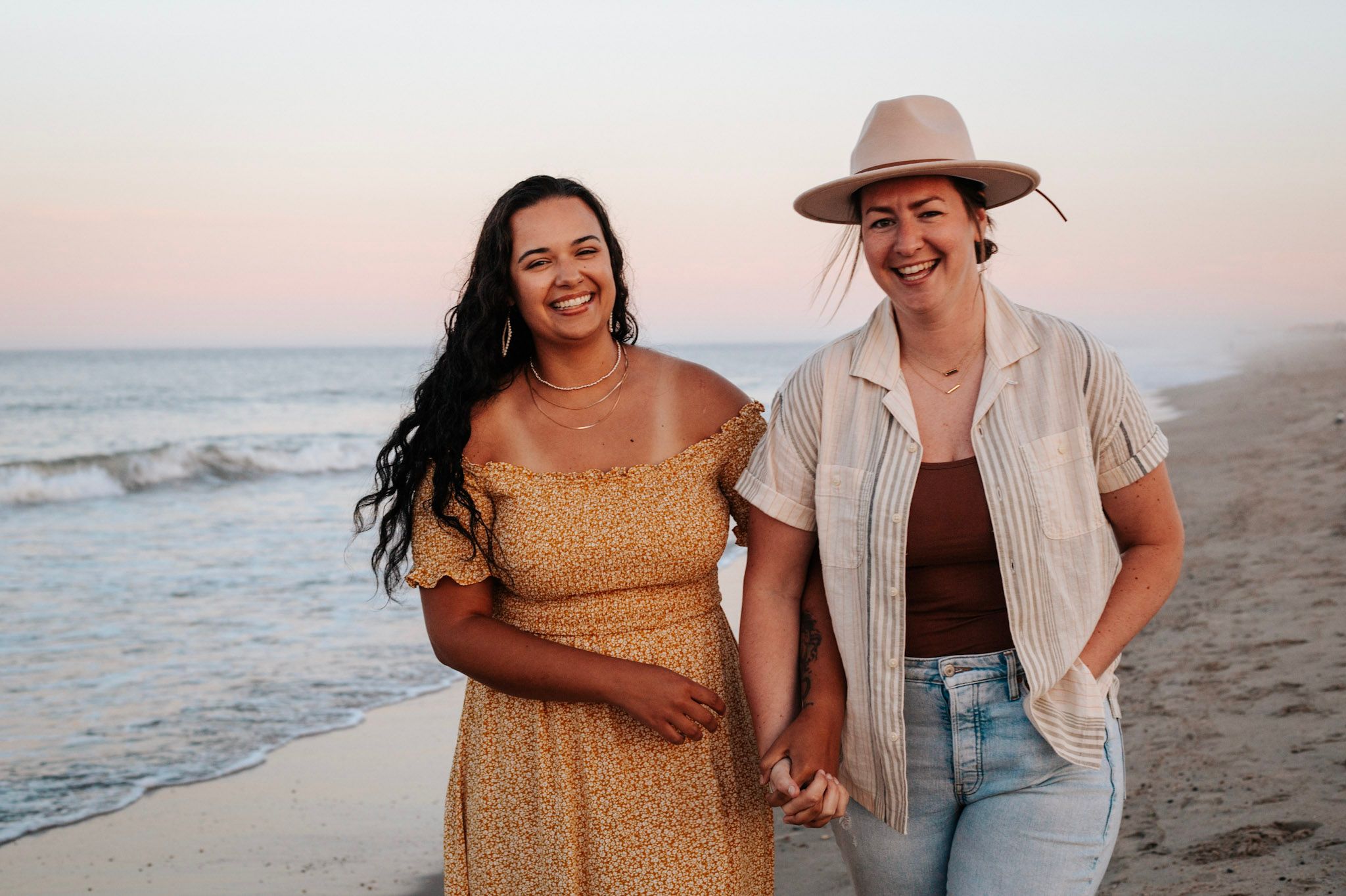 Two women smile and hold hands while walking on a beach at sunset, one in a floral dress and one in a hat.