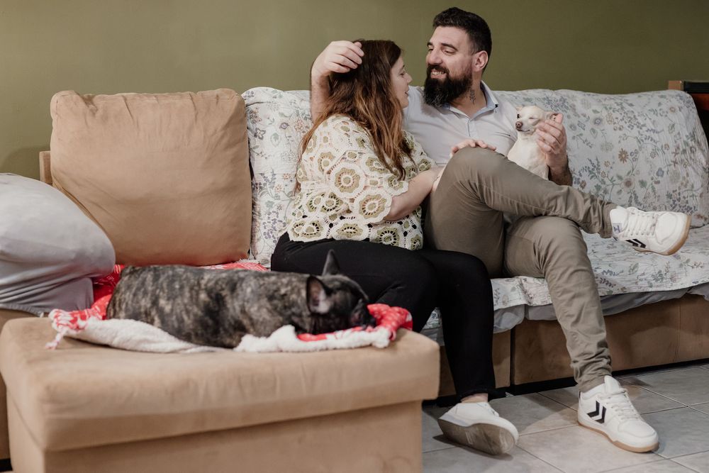 A person relaxes on a beige couch with a small white dog curled up beside them.