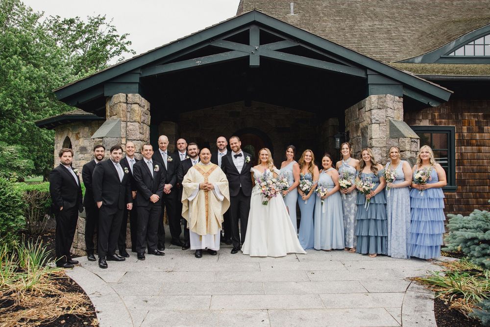Large wedding party in formal attire gathered outside stone church entrance with priest in white robes.