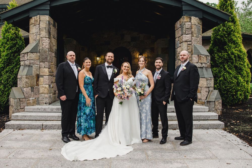 Wedding party stands together under stone archways at an elegant venue during outdoor ceremony.