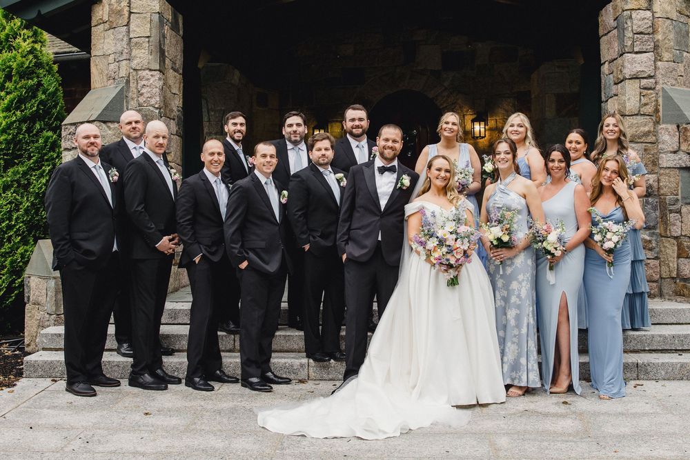 Wedding party in formal attire poses on stone steps with bridesmaids in blue dresses and groomsmen in black tuxedos.