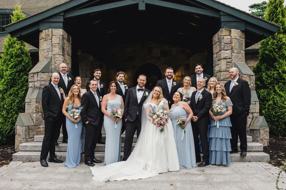 Wedding party poses together under stone pavilion in elegant formal attire with bridesmaids in light blue dresses.