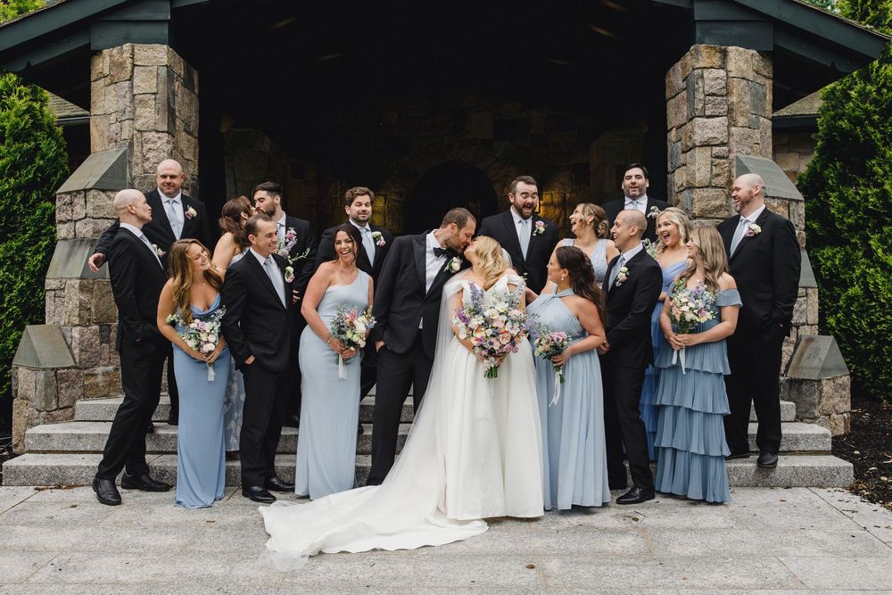 Wedding party in blue and gray attire poses together against stone wall backdrop.