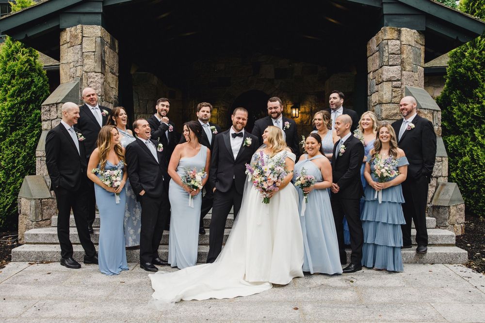 Wedding party in formal attire poses on lush green lawn with elegant stone building in background.