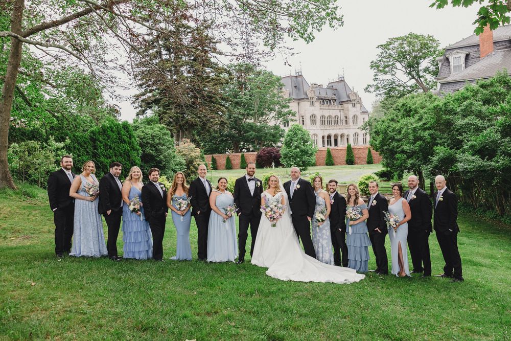 Wedding party stands in line on grassy lawn with historic stone mansion backdrop.