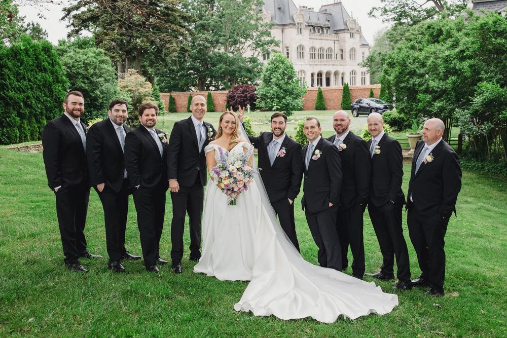 Elegant wedding party poses outdoors with classic stone architecture and landscaped gardens.