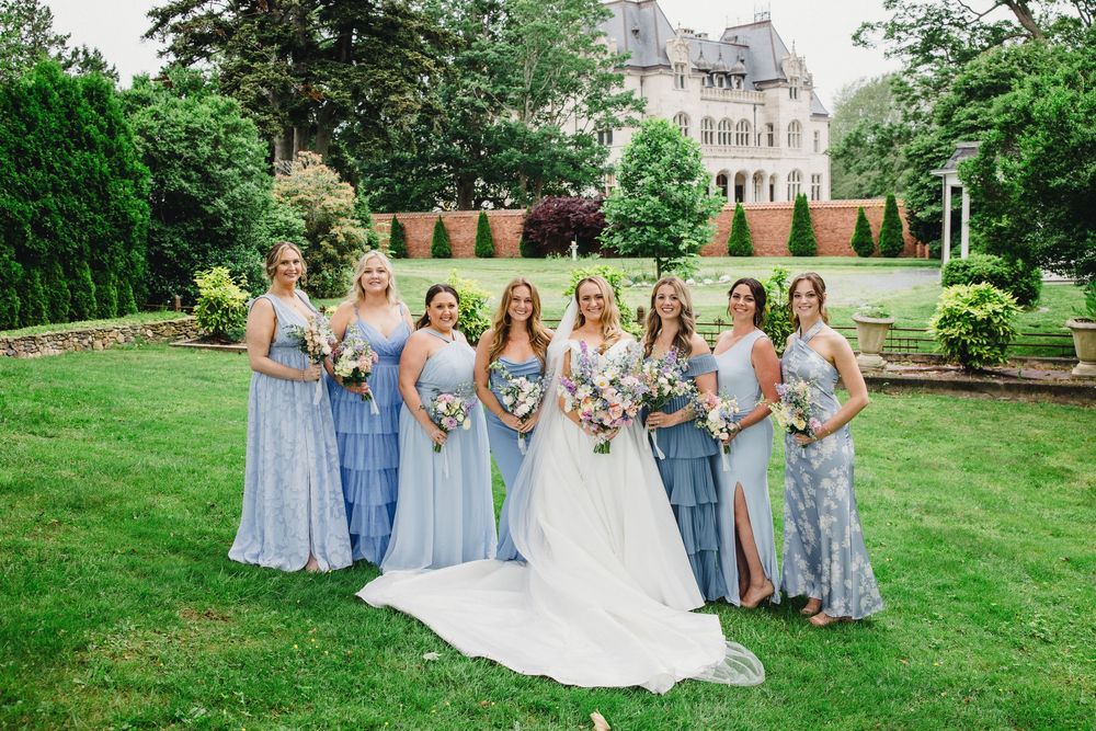 Wedding group in blue and black attire arranged on manicured lawn with historic building backdrop.