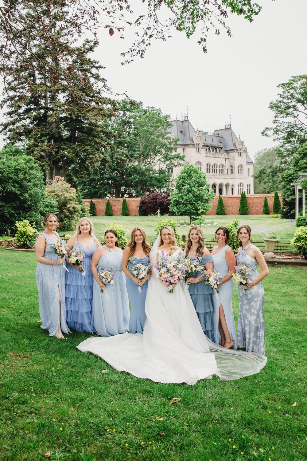 Wedding party in blue and gray dresses poses on a manicured lawn with a grand estate building in the background.