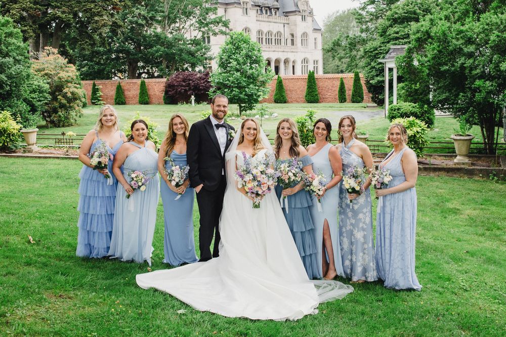 Wedding party with bridesmaids in light blue dresses poses on green lawn near stone mansion.