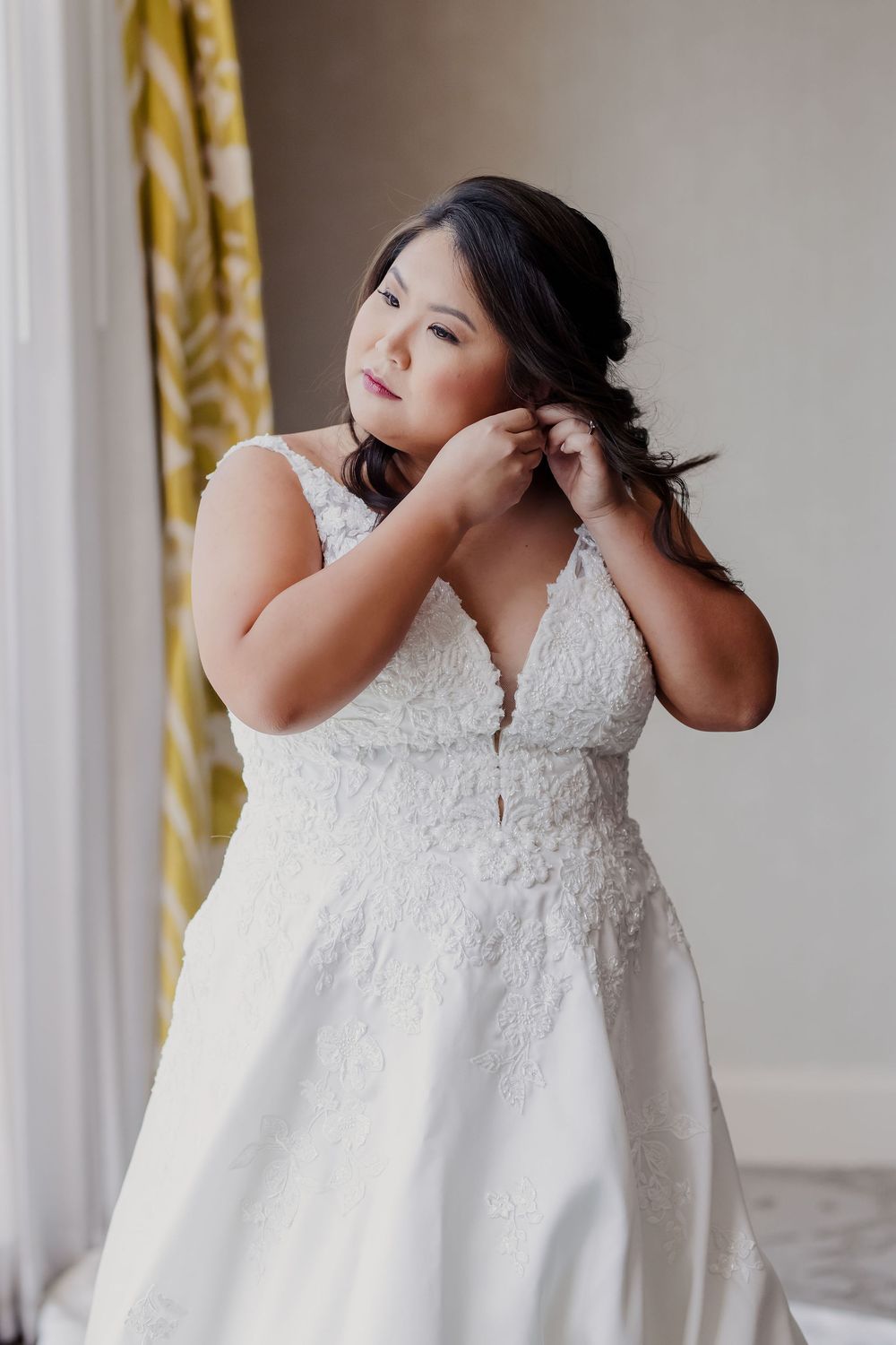 A wedding dress with intricate lace bodice and flowing white fabric hangs elegantly against a light wall.