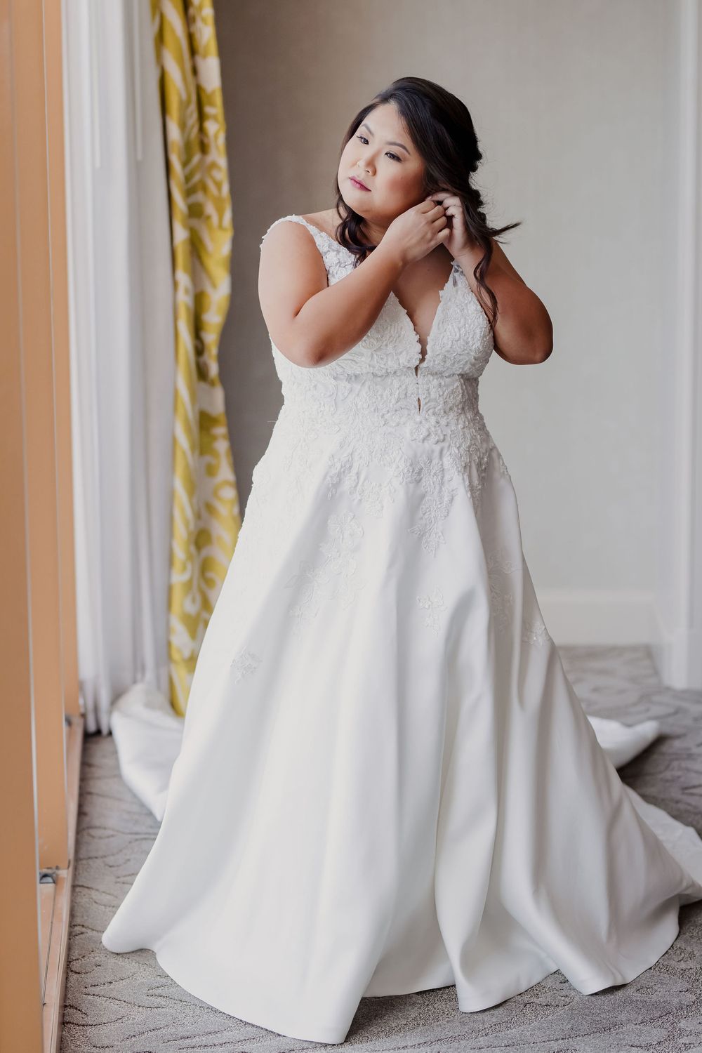 Plus size bride in a flowing white lace wedding gown with plunging neckline adjusts her hair near a window.