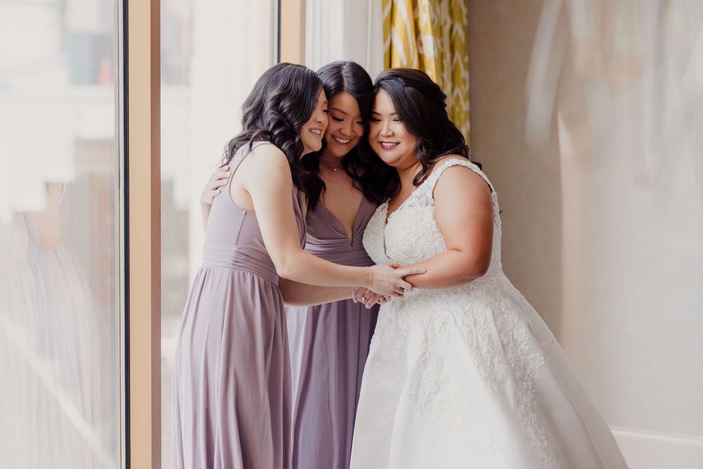 Bridesmaids in lavender dresses share a joyful moment with bride in white gown near bright window.