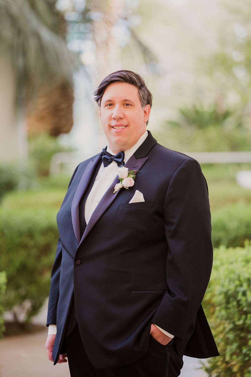 Groom in classic black tuxedo poses outdoors for formal wedding portraits with natural greenery in background.