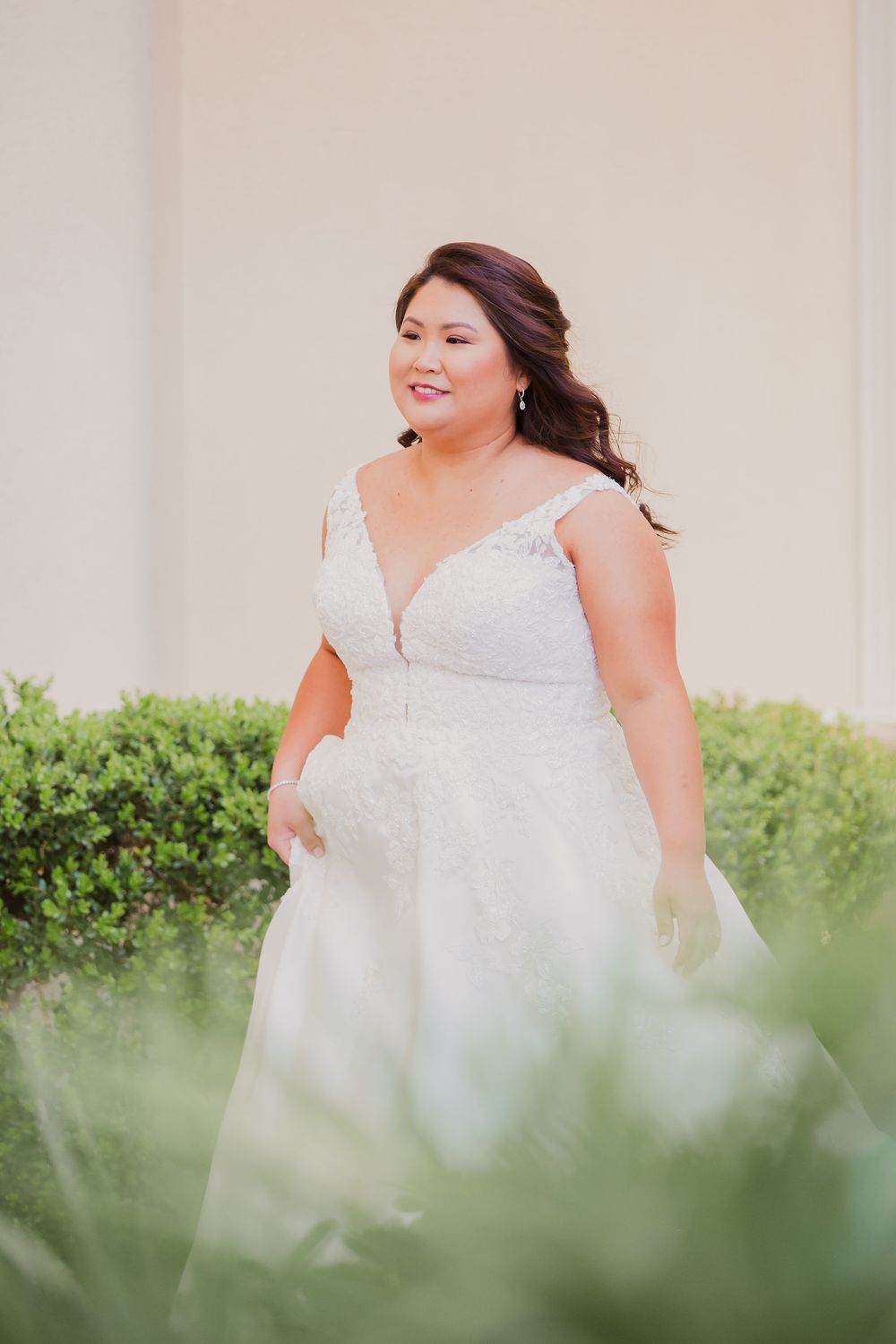 A bride in a white v-neck wedding gown stands among greenery in an outdoor garden setting.