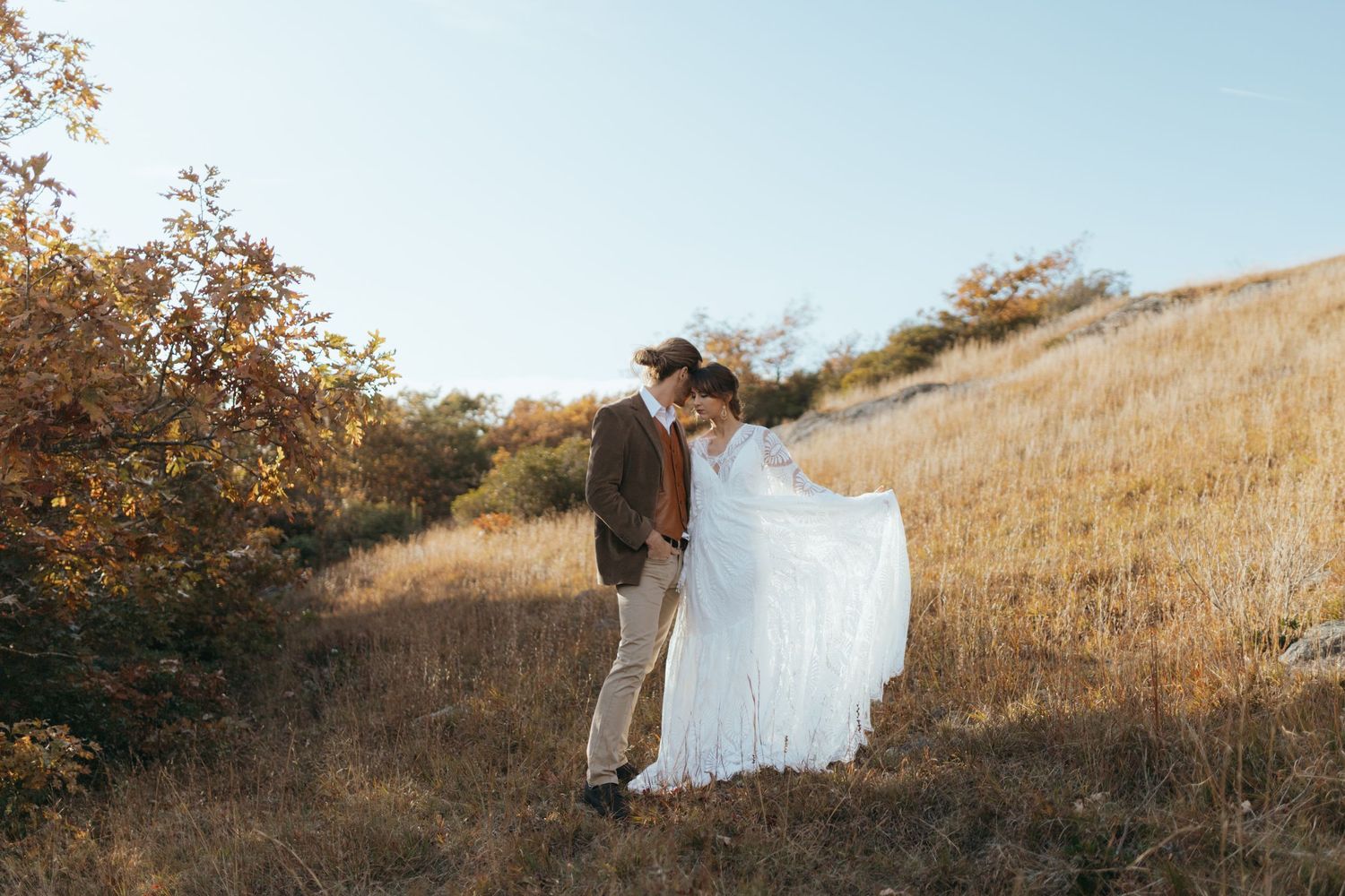 A couple in wedding attire stands together on a golden hillside during sunset.