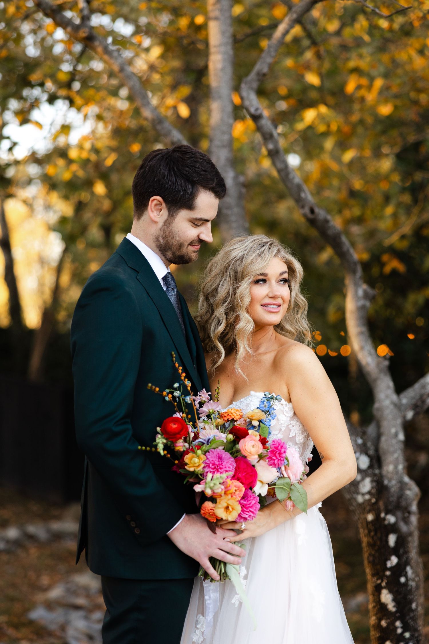 A couple in wedding attire stands among autumn trees with a colorful bouquet featuring bright pink and red flowers.