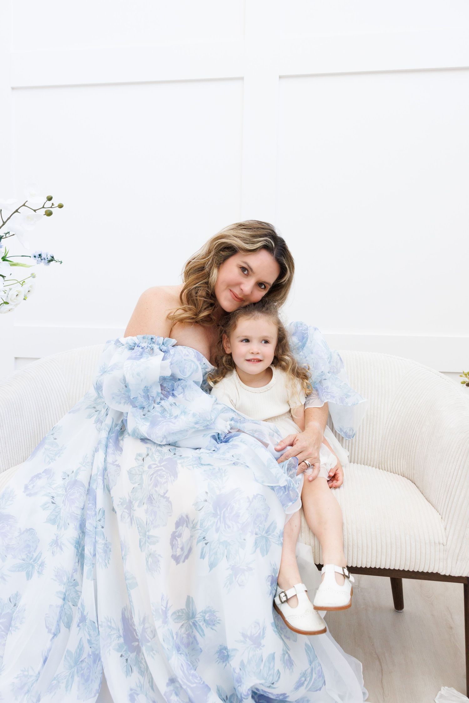 Mother and daughter in matching blue floral dresses sit together on a white couch in a bright room.