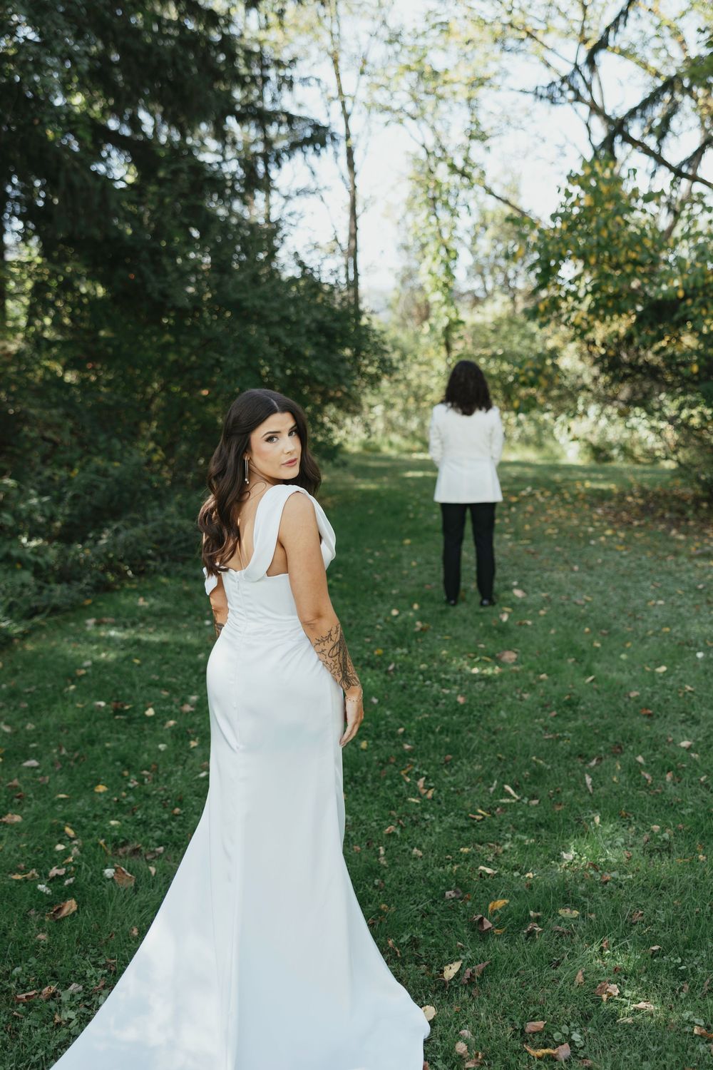 A bride in a flowing white dress twirls on a grassy lawn surrounded by autumn trees.