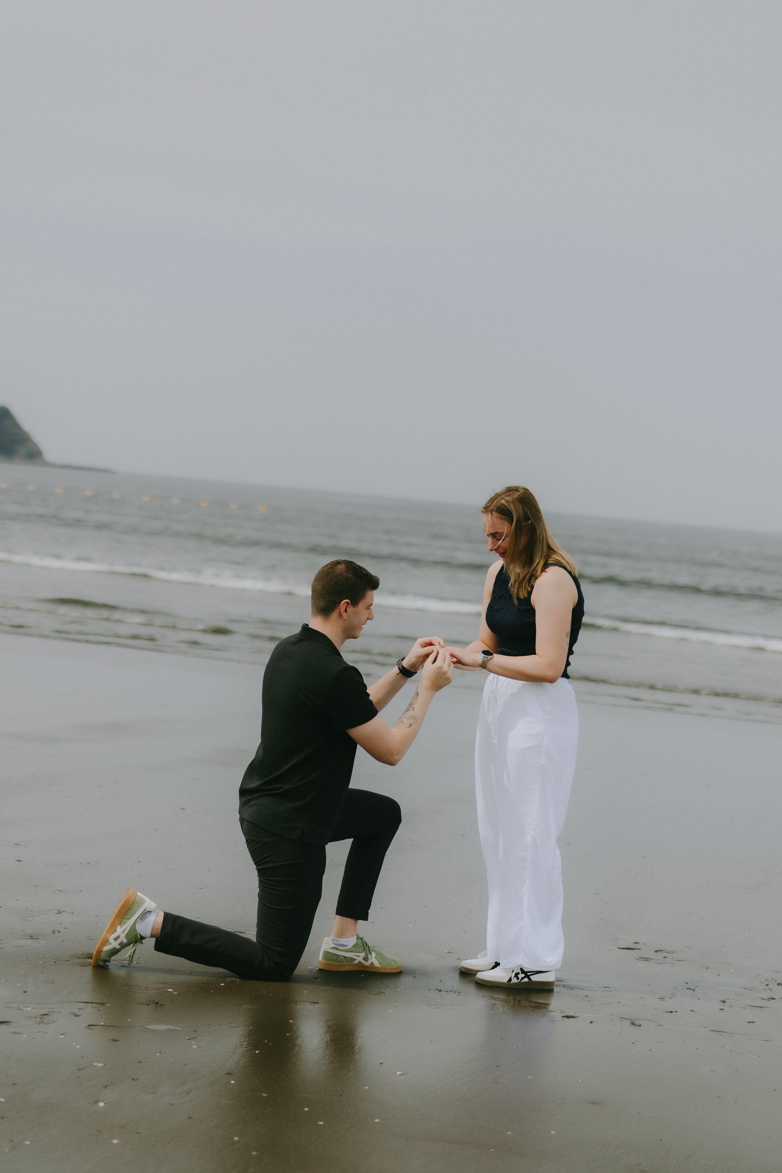 Man proposing on one knee to woman on a misty beach, holding her hand with the ocean in the background.