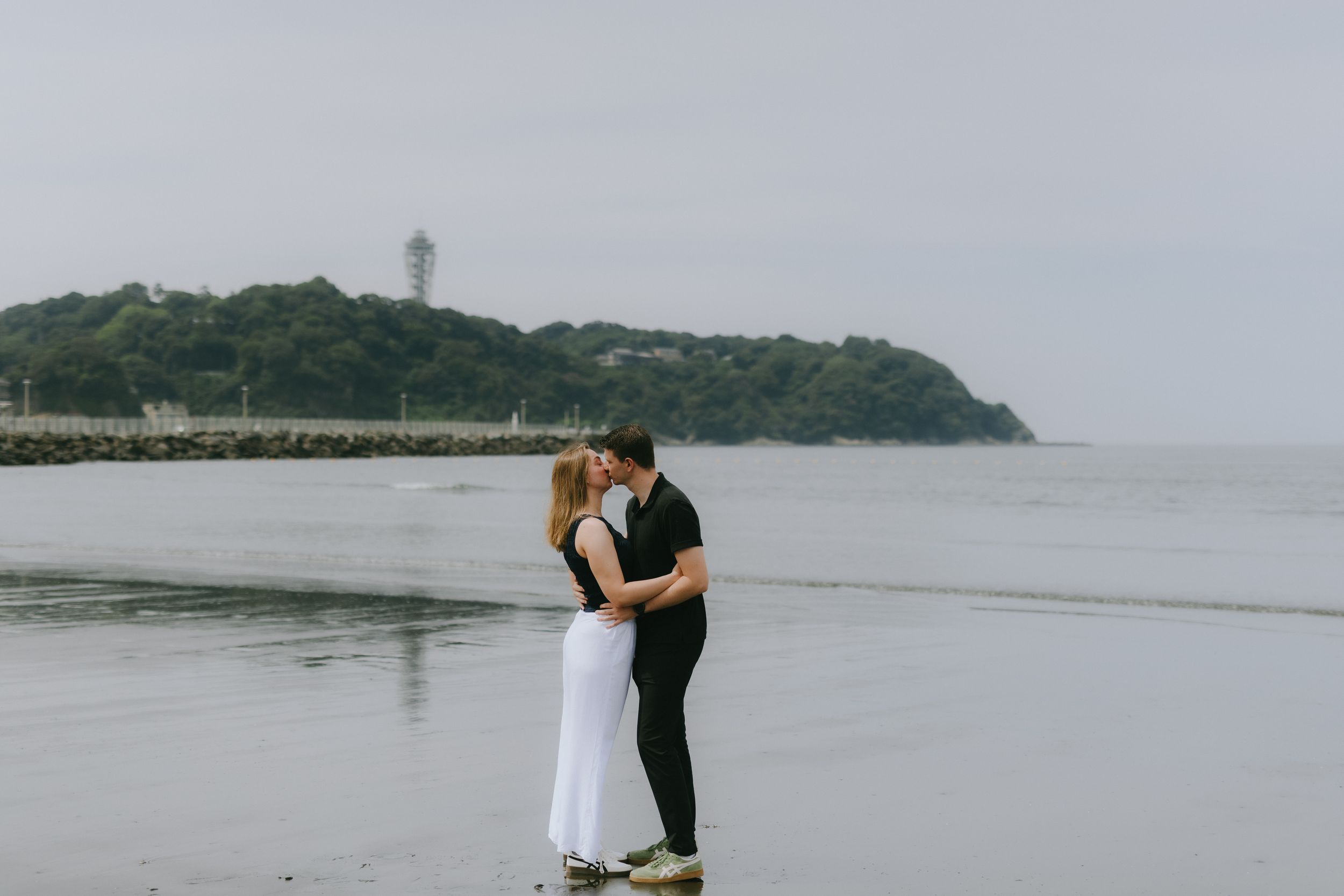 Couple sharing a romantic kiss on a misty beach with a lighthouse and lush green headland in the background.