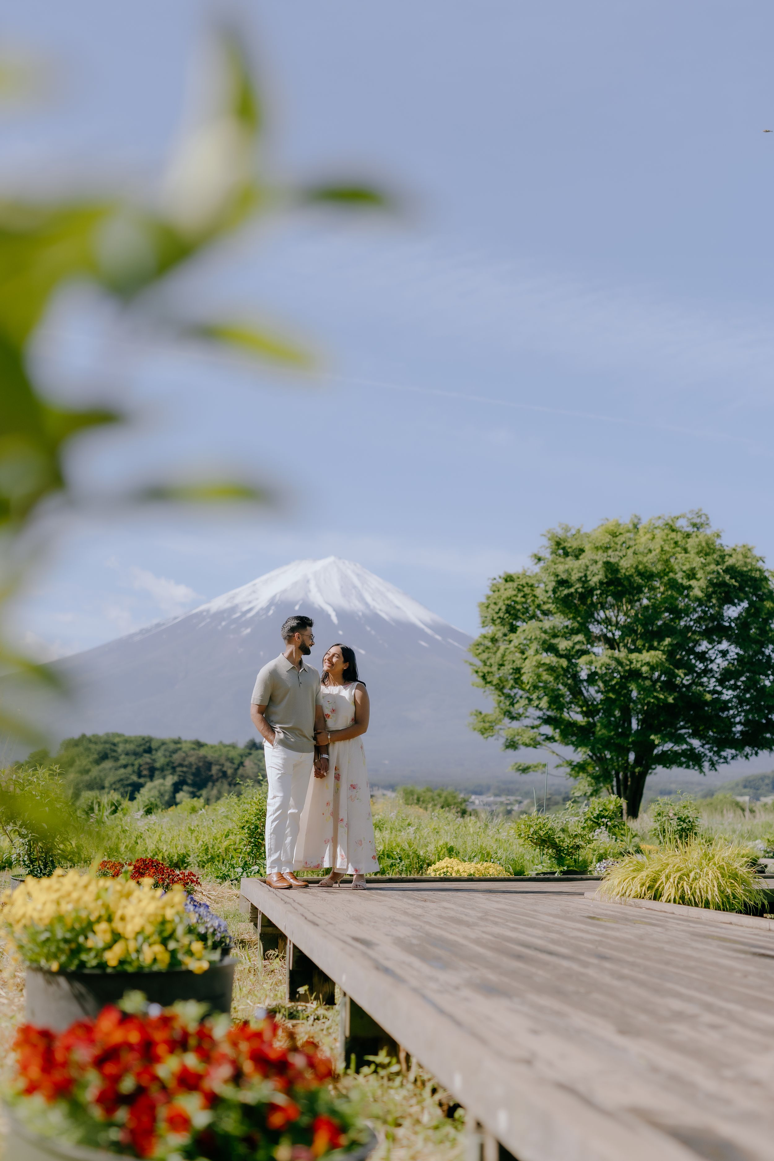 A couple stands on a wooden deck overlooking Mount Fuji in Japan with colorful flowers in the foreground.